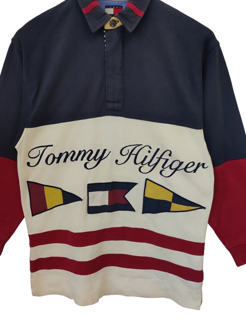 May include: A navy blue, red, and cream-colored long-sleeve shirt with the Tommy Hilfiger logo and nautical flag graphics. The shirt has a collared neckline and a buttoned placket.