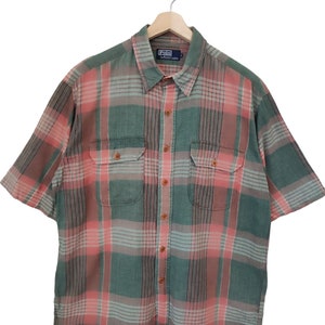 Peut inclure: Chemise à carreaux à manches courtes dans les tons de vert, rose et gris, avec deux poches de poitrine et des boutons en bois. La chemise est suspendue à un cintre en bois. Le logo Polo by Ralph Lauren est visible.