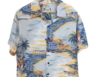 May include: Short-sleeved button-down shirt with a tropical landscape design. The shirt features a light blue background with yellow, blue, and green accents, depicting palm trees, buildings, and a beach scene. The shirt is hanging on a wooden hanger.
