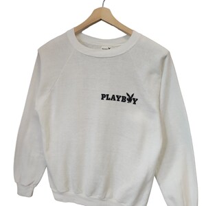 May include: White sweatshirt with the text "PLAYBOY" in black, along with the iconic bunny logo. The long-sleeved top is hanging on a wooden hanger. The sweatshirt has a crew neck and raglan sleeves.