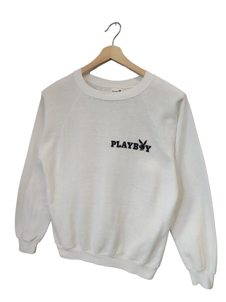 May include: White sweatshirt with the text "PLAYBOY" in black, along with the iconic bunny logo. The long-sleeved top is hanging on a wooden hanger. The sweatshirt has a crew neck and raglan sleeves.