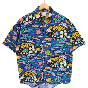 Könnte beinhalten: Kurzärmeliges Hemd mit einem lebendigen, farbenfrohen Muster. Das blaue Hemd zeigt ein sich wiederholendes Design aus Fischen, Sternen und abstrakten Formen in Gelb, Rosa, Grün und Lila. Es hängt an einem Holzbügel.