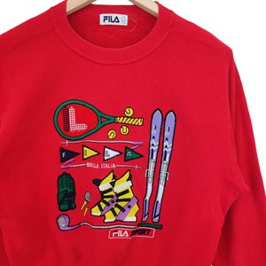 May include: Red FILA sweatshirt with embroidered sports graphics, including a tennis racket, skis, and golf club. The sweatshirt features the text "BIELLA ITALIA" and "FILA SPORT".