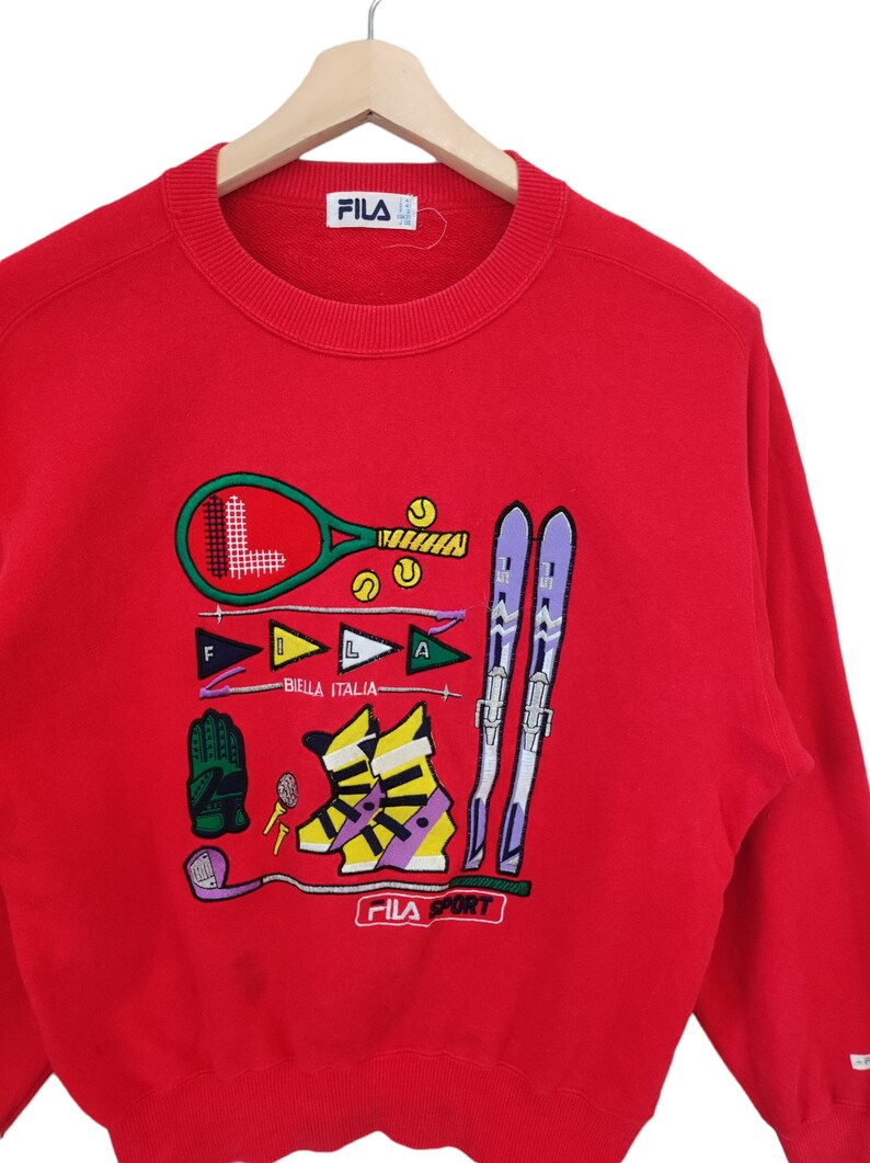 May include: Red FILA sweatshirt with embroidered sports graphics, including a tennis racket, skis, and golf club. The sweatshirt features the text "BIELLA ITALIA" and "FILA SPORT".