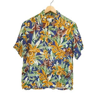 May include: A short-sleeved button-down shirt with a vibrant floral and tiger print. The shirt features a blue background with yellow flowers, green leaves, and brown tigers. The shirt is hanging on a wooden hanger.