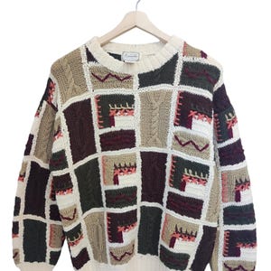 Könnte beinhalten: Ein Vintage-Patchwork-Pullover in neutralen Tönen, darunter Beige, Creme, Braun und Burgunderrot. Der Pullover hat einen Rundhalsausschnitt und lange Ärmel mit einer Vielzahl von strukturierten Strickquadraten und dekorativen Nähten. Der Pullover wird auf einem Holzbügel präsentiert.