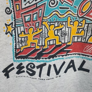 May include: Light gray sweatshirt with a colorful graphic design. The design includes the word "ART" and "FESTIVAL" in bold letters, along with yellow figures, buildings, and abstract shapes. The text "CAMELFLAUGE HARD WEAR '92" is also visible.