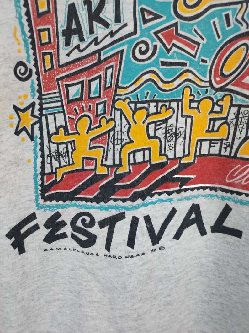 May include: Light gray sweatshirt with a colorful graphic design. The design includes the word "ART" and "FESTIVAL" in bold letters, along with yellow figures, buildings, and abstract shapes. The text "CAMELFLAUGE HARD WEAR '92" is also visible.