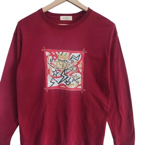 Puede incluir: Una camiseta de manga larga de color burdeos con un dise&ntilde;o gr&aacute;fico. El dise&ntilde;o presenta la palabra "THOR" y varias formas y estrellas dentro de un cuadrado con l&iacute;neas rojas. La camiseta est&aacute; colgada en una percha de madera.