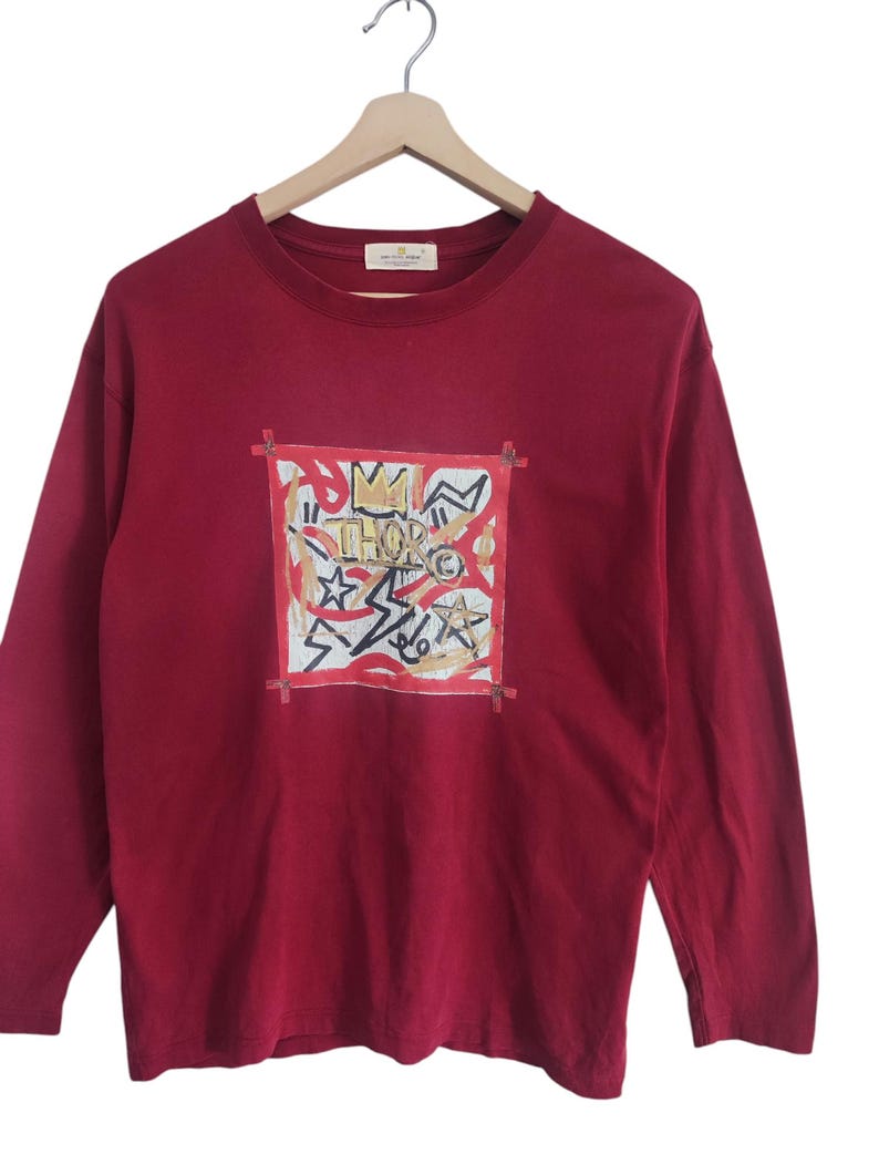 Puede incluir: Una camiseta de manga larga de color burdeos con un dise&ntilde;o gr&aacute;fico. El dise&ntilde;o presenta la palabra "THOR" y varias formas y estrellas dentro de un cuadrado con l&iacute;neas rojas. La camiseta est&aacute; colgada en una percha de madera.
