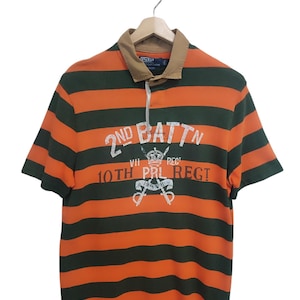 May include: Orange and green striped polo shirt with short sleeves. The shirt features a tan collar and the text "2ND BATTN 10TH PRL REGT" in white. The shirt is hanging on a wooden hanger.
