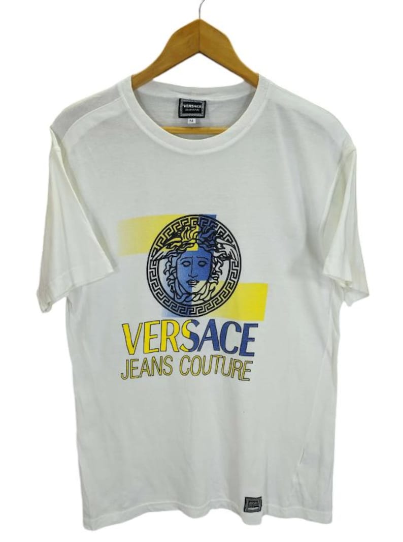 May include: White t-shirt with a graphic print featuring a Medusa head design in blue, yellow, and black. The text "VERSACE JEANS COUTURE" is printed below the graphic. The shirt is hanging on a wooden hanger.
