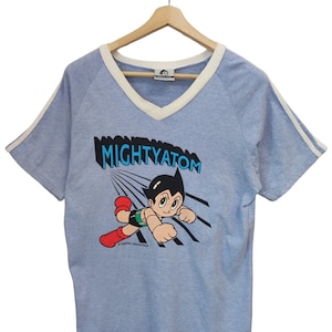 May include: Light blue t-shirt with a white V-neck and sleeve trim. The shirt features a graphic of the character Astro Boy with the text "MIGHTY ATOM". The shirt is displayed on a wooden hanger.