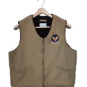 May include: Tan vest with a black interior lining, a full-length zipper, and two front pockets. The vest features a patch on the chest with a winged emblem and the text "ARMY AIR FORCES". The vest is hanging on a wooden hanger.