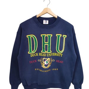 May include: A navy blue crewneck sweatshirt featuring the text DHU and DUCK HEAD UNIVERSITY in green and yellow lettering. Below, a crest logo with a duck graphic is flanked by the words DUCK and HEAD, with ESTABLISHED 1865 printed below.