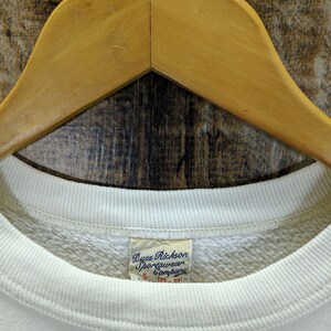 May include: Cream-coloured Buzz Rickson Sportswear Company sweatshirt, size S (36-38), hanging on a wooden hanger. The label is visible, and the sweatshirt has a ribbed neckline and cuffs. The background is a textured, wooden surface.