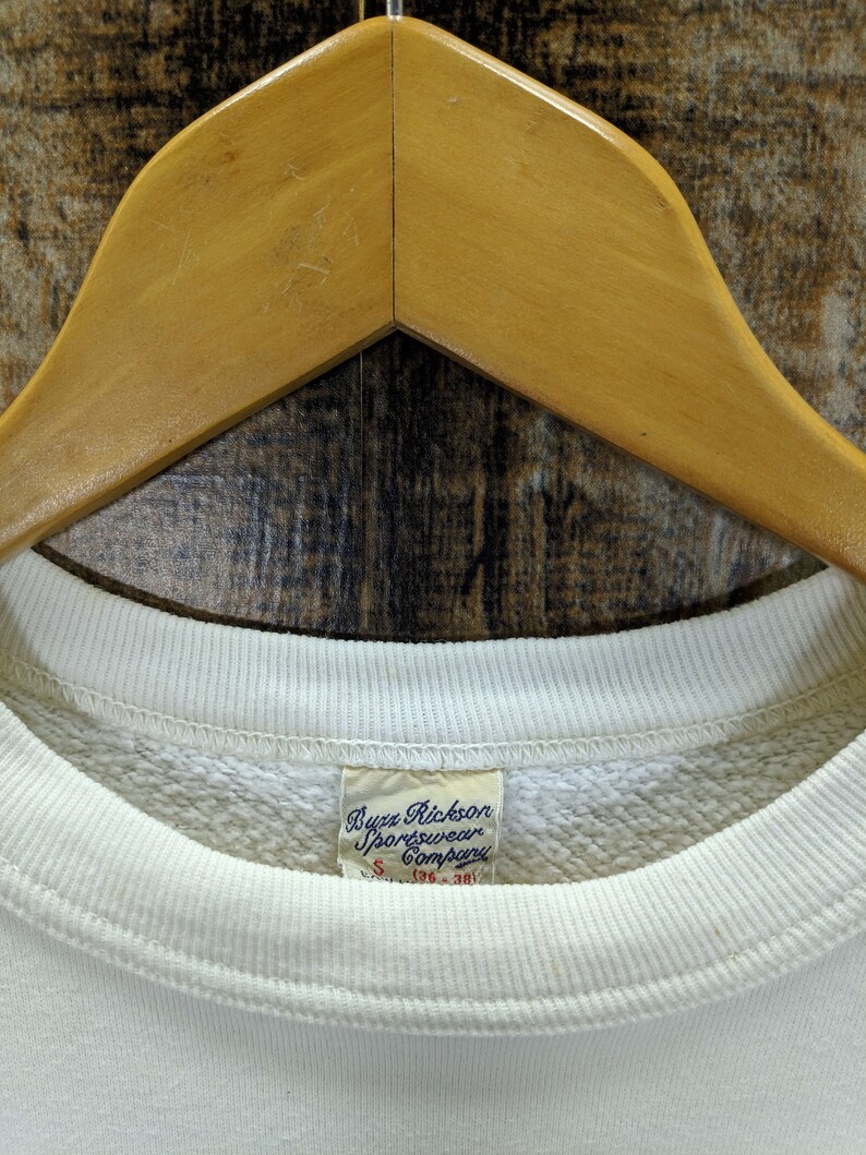 May include: Cream-coloured Buzz Rickson Sportswear Company sweatshirt, size S (36-38), hanging on a wooden hanger. The label is visible, and the sweatshirt has a ribbed neckline and cuffs. The background is a textured, wooden surface.