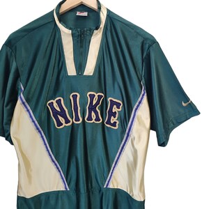 May include: Green and cream-colored Nike athletic shirt with a zip-up collar. The shirt features the Nike logo in navy blue with a gold outline across the chest. A small Nike swoosh is visible on the sleeve.