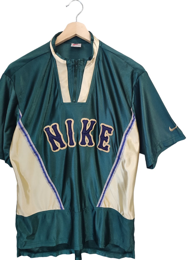 May include: Green and cream-colored Nike athletic shirt with a zip-up collar. The shirt features the Nike logo in navy blue with a gold outline across the chest. A small Nike swoosh is visible on the sleeve.