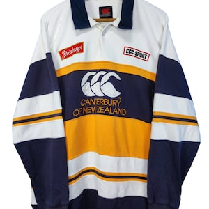 May include: A long-sleeved rugby shirt in white, navy blue, and gold. The shirt features the "Canterbury of New Zealand" logo, along with "Steinlager" and "CCC SPORT" patches. The shirt is hanging on a wooden hanger.