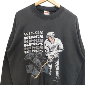 K&ouml;nnte beinhalten: Dunkelgraues Vintage-Sweatshirt mit einer Hockey-Spieler-Grafik und dem Wort "KINGS" in Wei&szlig;. Das Sweatshirt hat einen Rundhalsausschnitt und lange &Auml;rmel. Ein Mitchell & Ness-Etikett ist sichtbar.
