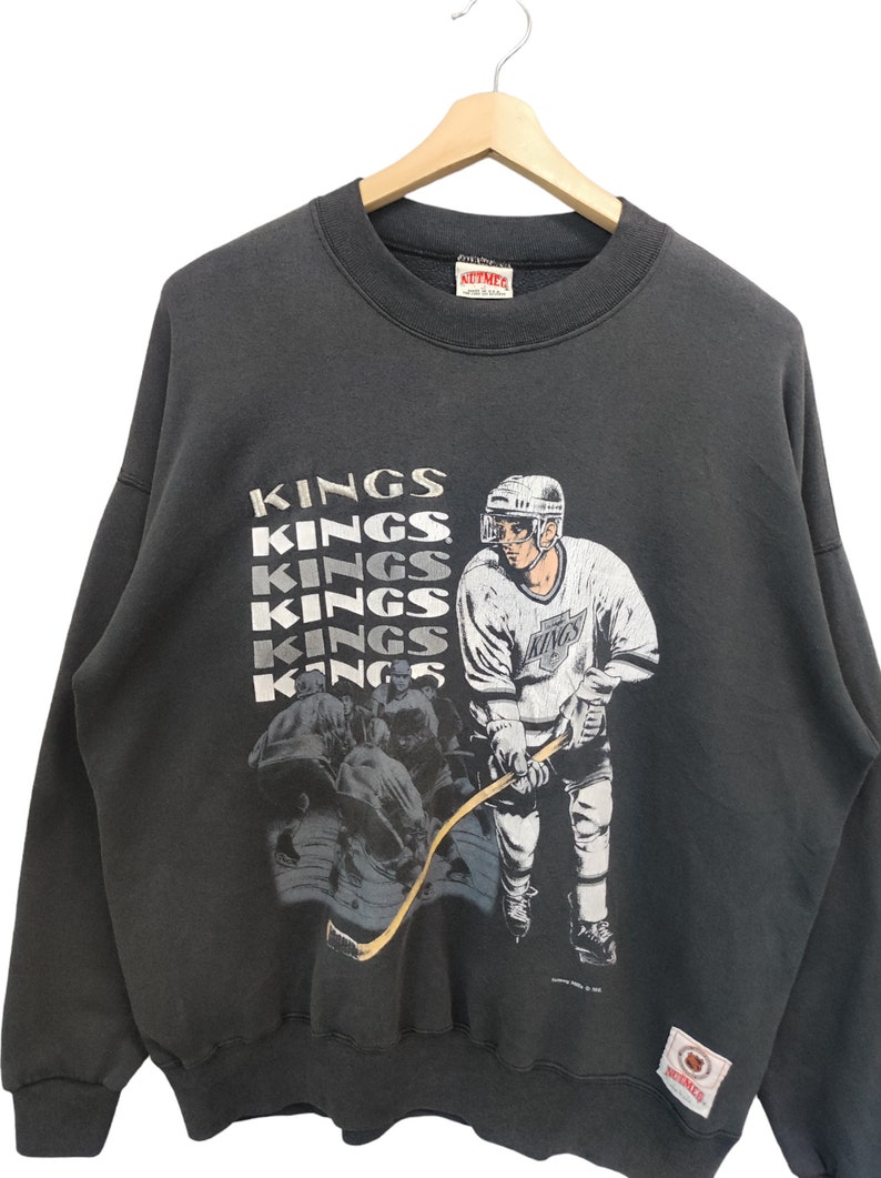 K&ouml;nnte beinhalten: Dunkelgraues Vintage-Sweatshirt mit einer Hockey-Spieler-Grafik und dem Wort "KINGS" in Wei&szlig;. Das Sweatshirt hat einen Rundhalsausschnitt und lange &Auml;rmel. Ein Mitchell & Ness-Etikett ist sichtbar.