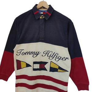 May include: A long-sleeved rugby shirt in navy, red, and cream, featuring the Tommy Hilfiger logo and nautical flag graphics. The shirt has a collared neckline with a button closure and red accents.