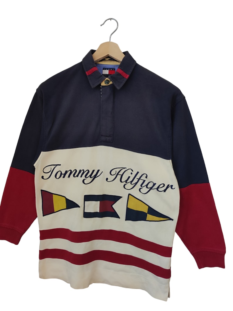 May include: A long-sleeved rugby shirt in navy, red, and cream, featuring the Tommy Hilfiger logo and nautical flag graphics. The shirt has a collared neckline with a button closure and red accents.
