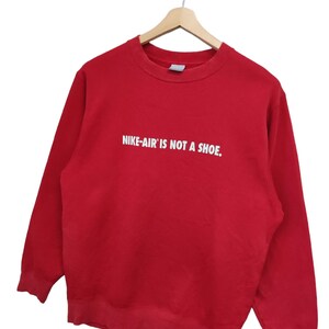 May include: A red crew neck sweatshirt with the text "NIKE-AIR IS NOT A SHOE." in white block letters. The sweatshirt is hanging on a wooden hanger against a white background. A small Nike logo is visible near the bottom hem.