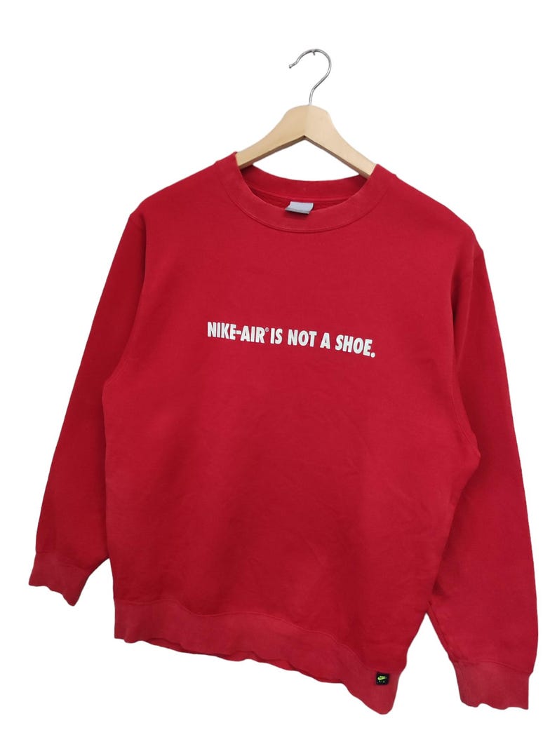 May include: A red crew neck sweatshirt with the text "NIKE-AIR IS NOT A SHOE." in white block letters. The sweatshirt is hanging on a wooden hanger against a white background. A small Nike logo is visible near the bottom hem.