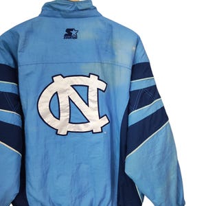 May include: A light blue and navy blue Starter brand windbreaker jacket featuring a large white embroidered North Carolina Tar Heels logo on the back. The jacket has a high collar, long sleeves with navy blue panels, and a ribbed hem.
