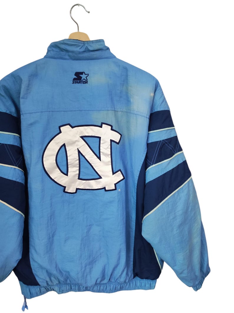 May include: A light blue and navy blue Starter brand windbreaker jacket featuring a large white embroidered North Carolina Tar Heels logo on the back. The jacket has a high collar, long sleeves with navy blue panels, and a ribbed hem.