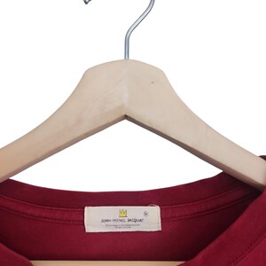 Puede incluir: Una camiseta granate cuelga de una percha de madera. La camiseta tiene una etiqueta con el texto "JEAN-MICHEL BASQUIAT" y un logotipo de corona. La camiseta es de color liso y parece ser de algod&oacute;n.