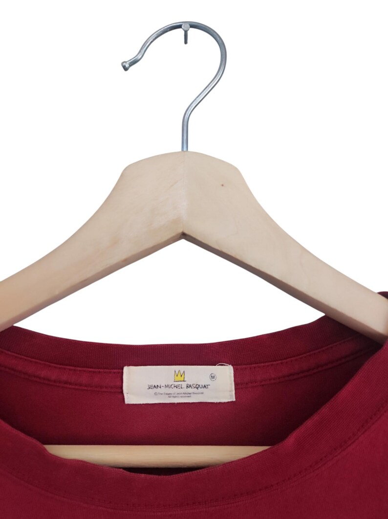 Puede incluir: Una camiseta granate cuelga de una percha de madera. La camiseta tiene una etiqueta con el texto "JEAN-MICHEL BASQUIAT" y un logotipo de corona. La camiseta es de color liso y parece ser de algod&oacute;n.