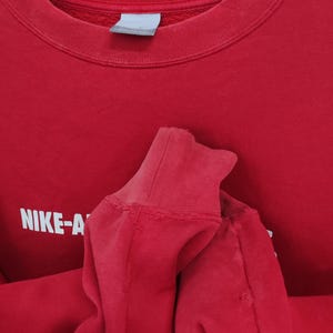 May include: A red crew neck sweatshirt with the text "NIKE-A" in white block letters. The sweatshirt has a ribbed collar and cuffs. The image is a close-up of the garment, showing the texture of the fabric.