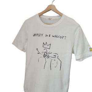 Puede incluir: Camiseta blanca con arte gr&aacute;fico y texto en negro. El texto "JERSEY JOE WALCOTT" est&aacute; encima de un dibujo de dos figuras y una corona. Una peque&ntilde;a corona est&aacute; en la manga. La camiseta est&aacute; colgada en una percha de madera.