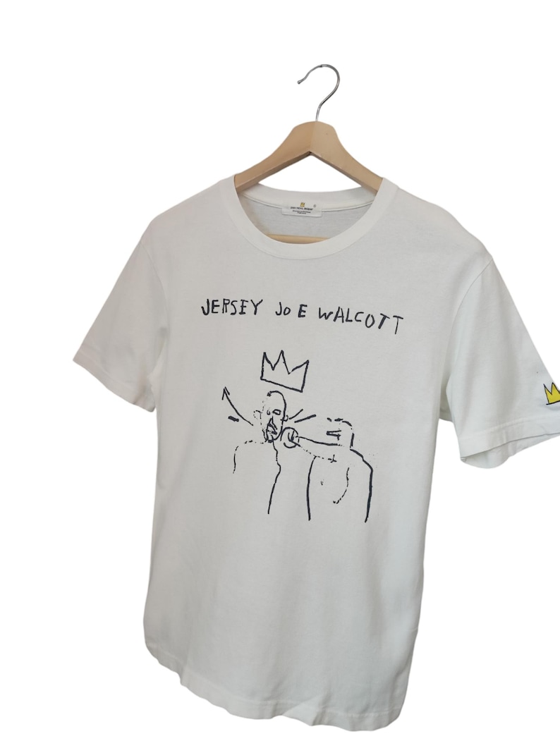 Puede incluir: Camiseta blanca con arte gr&aacute;fico y texto en negro. El texto "JERSEY JOE WALCOTT" est&aacute; encima de un dibujo de dos figuras y una corona. Una peque&ntilde;a corona est&aacute; en la manga. La camiseta est&aacute; colgada en una percha de madera.