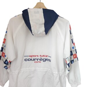 May include: White Courr&egrave;ges jacket with a navy blue hood and trim. The jacket features geometric patterns in red, blue, and gray on the sleeves. The text "sport futur courr&egrave;ges paris" is printed on the back.