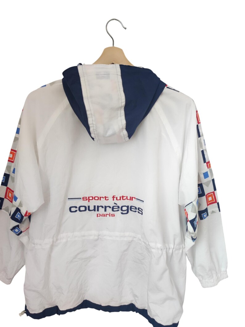 May include: White Courr&egrave;ges jacket with a navy blue hood and trim. The jacket features geometric patterns in red, blue, and gray on the sleeves. The text "sport futur courr&egrave;ges paris" is printed on the back.