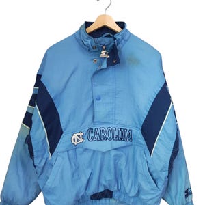 May include: A light blue and navy blue pullover jacket featuring the text CAROLINA and a logo on the front. The jacket has a high collar, a partial front zip with a Starter brand pull, snap button closures, and long sleeves with elastic cuffs.