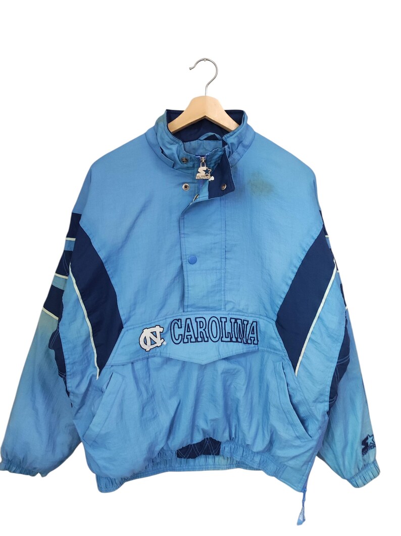 May include: A light blue and navy blue pullover jacket featuring the text CAROLINA and a logo on the front. The jacket has a high collar, a partial front zip with a Starter brand pull, snap button closures, and long sleeves with elastic cuffs.