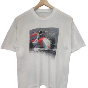 May include: White t-shirt featuring a graphic of a Formula 1 race car in red, white, and gray. The car has the number 7 on it. The shirt is hanging on a wooden hanger.