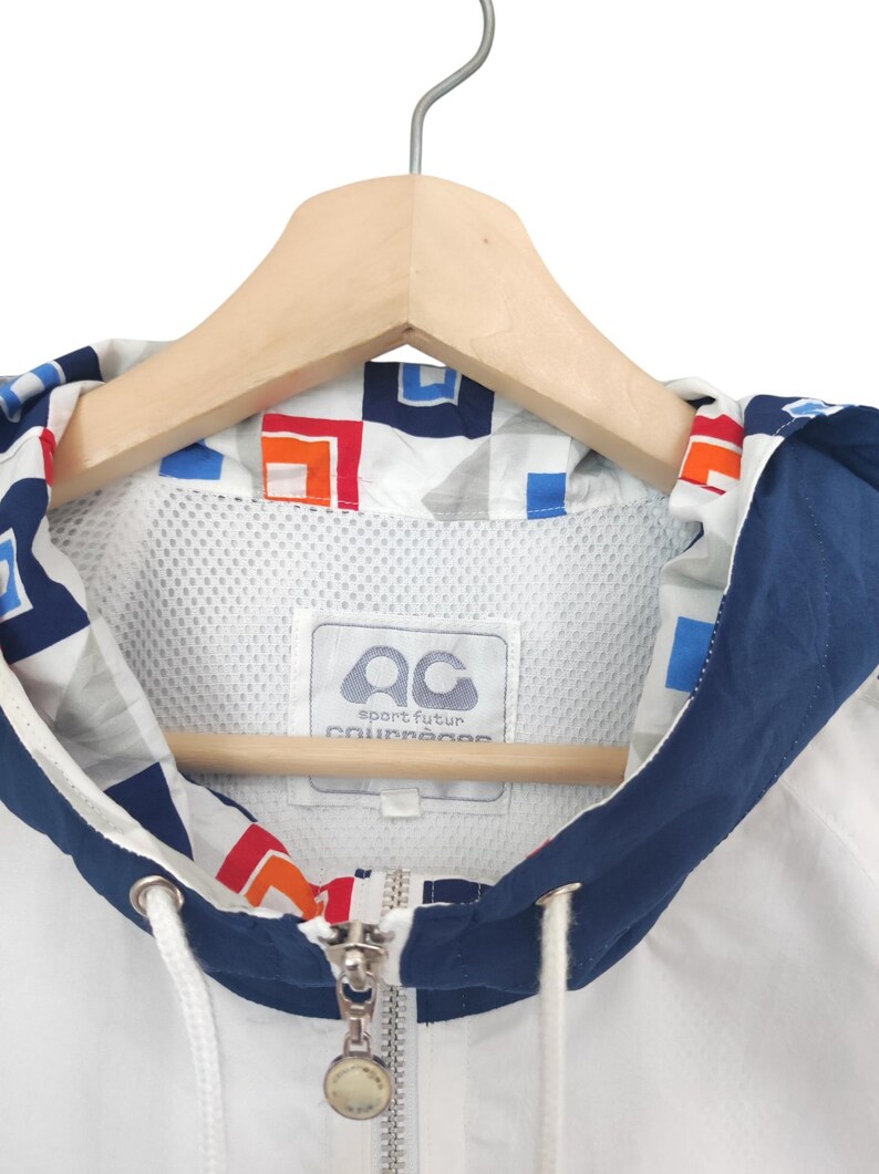 May include: White and navy blue hooded jacket with a geometric pattern on the hood lining. The jacket features a full zipper, drawstring, and a label with the text "sport futur collection". The jacket is hanging on a wooden hanger.