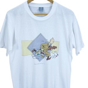 May include: White t-shirt featuring a cartoon graphic of a man in a suit, a white dog, and a suitcase. The graphic is set within a blue diamond and yellow rectangle. The shirt is hanging on a wooden hanger.