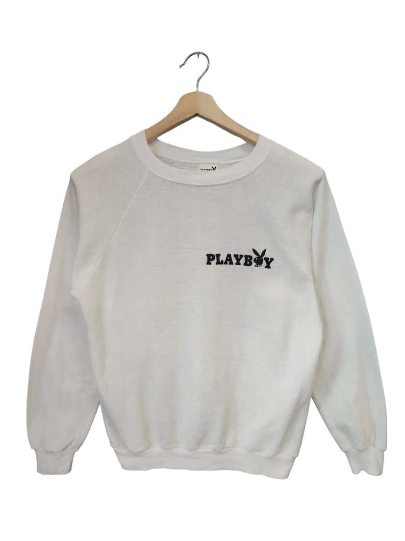 May include: Off-white sweatshirt with long sleeves, hanging on a wooden hanger. The black text "PLAYBOY" and a bunny logo are printed on the front. The sweatshirt is made of a soft material.