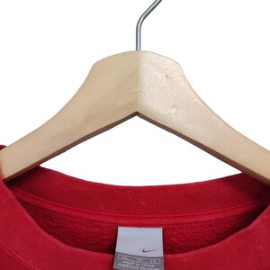 May include: A red crew neck sweatshirt hanging on a wooden hanger. The sweatshirt has a ribbed collar and a tag that reads "MADE IN CHINA" and "FABRIQUE EN CHINE". The Nike logo is visible on the tag.