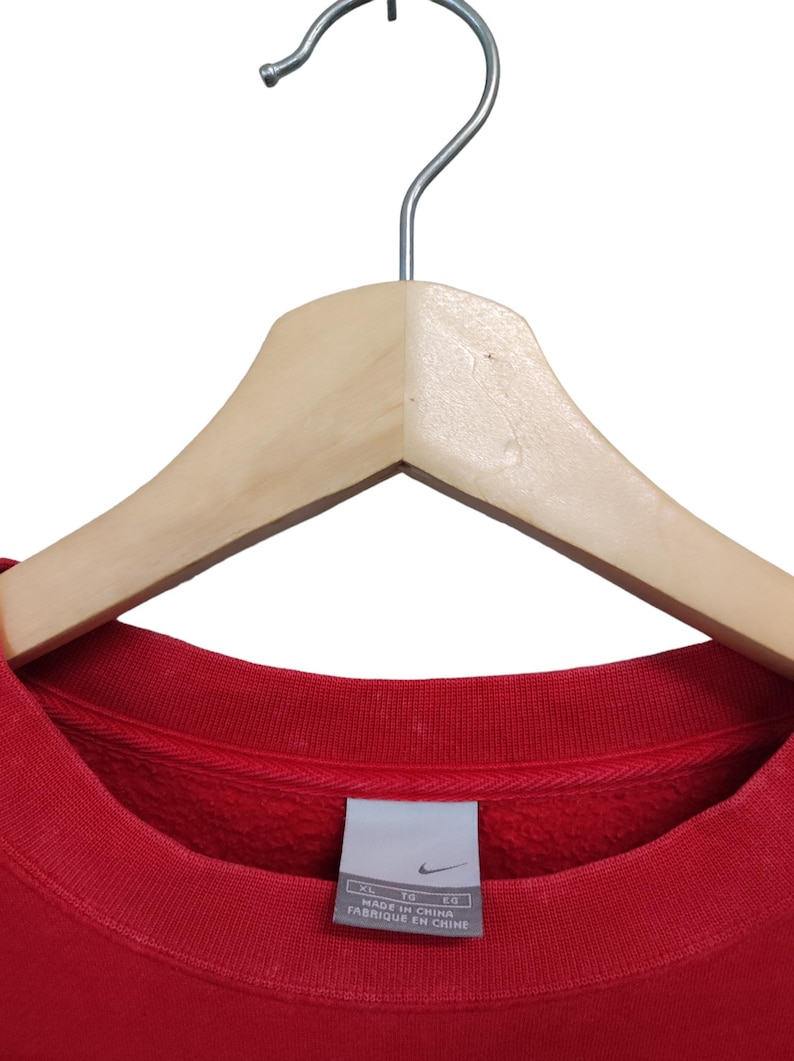 May include: A red crew neck sweatshirt hanging on a wooden hanger. The sweatshirt has a ribbed collar and a tag that reads "MADE IN CHINA" and "FABRIQUE EN CHINE". The Nike logo is visible on the tag.
