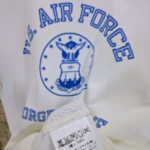 May include: Cream-coloured sweatshirt featuring the words "U.S. AIR FORCE" in blue, and a blue emblem. The label indicates it is 100% cotton. The text "ORG" is also visible. The label also includes Japanese text.