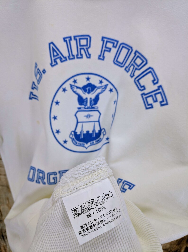 May include: Cream-coloured sweatshirt featuring the words "U.S. AIR FORCE" in blue, and a blue emblem. The label indicates it is 100% cotton. The text "ORG" is also visible. The label also includes Japanese text.