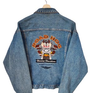 May include: Light blue denim jacket with a Road Hog design featuring the Tasmanian Devil riding a motorcycle. The back of the jacket displays the Harley Davidson Motorcycles logo. The jacket is hanging on a wooden hanger.